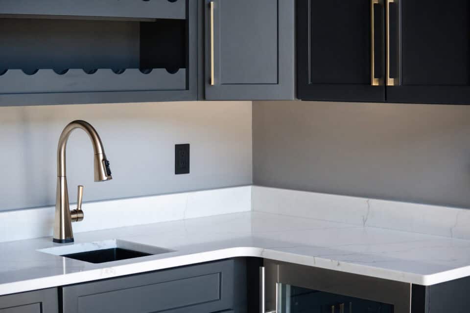 A modern kitchen corner in Knoxville features sleek dark grey cabinets, a natural stone countertop, and a stainless steel faucet. A built-in wine rack and under-cabinet lighting enhance the contemporary, elegant appearance.