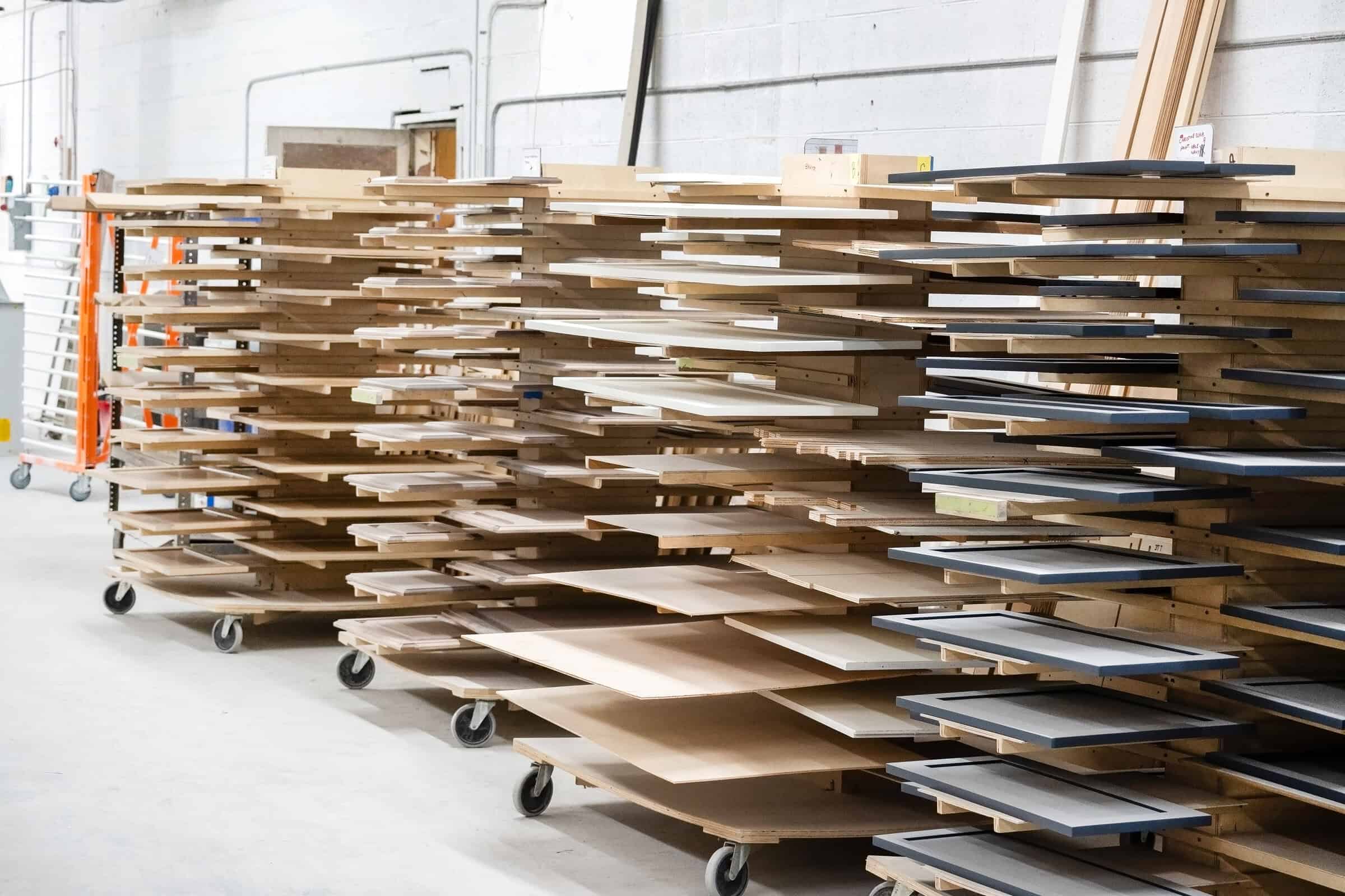 Stacks of wooden boards and panels are organized on wheeled racks in Tamco's workshop. Various sizes and shades of wood complement the scattered tools.