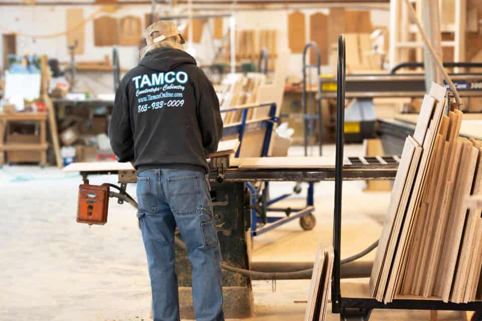 A person wearing a TAMCO hoodie operates a woodworking machine in a workshop filled with various wooden boards, tools, and cabinets.