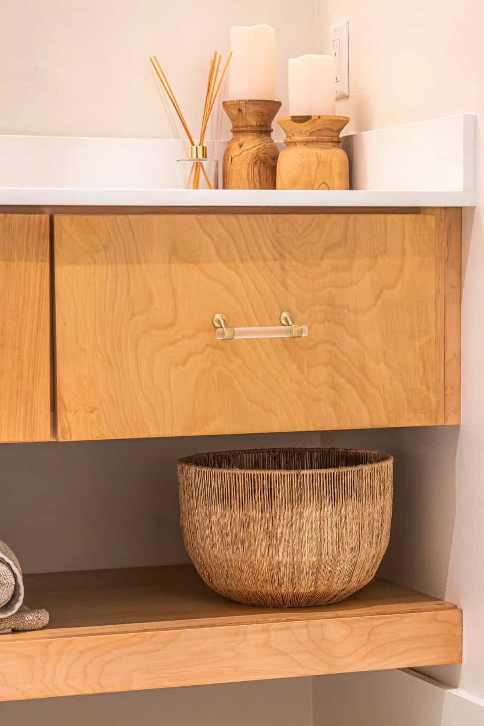 A modern wooden bathroom vanity with a quartz countertop, featuring a candle and reed diffuser. A textured basket rests on a lower shelf beside folded towels, enhancing the warm and natural aesthetic, while the sleek cabinets offer ample storage.