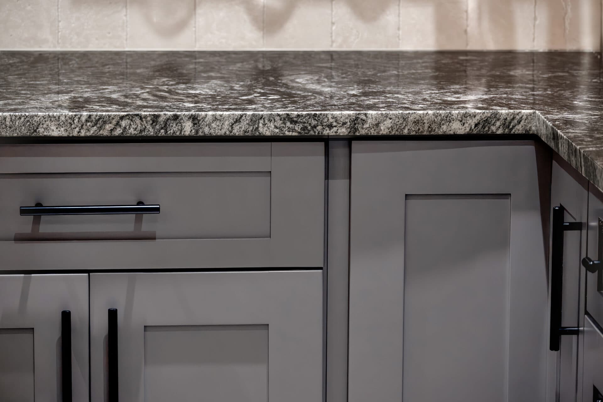 Gray kitchen cabinets with sleek black handles are topped with a natural stone countertop. The countertop features a dark and light speckled pattern, perfectly complementing the modern kitchen aesthetic.