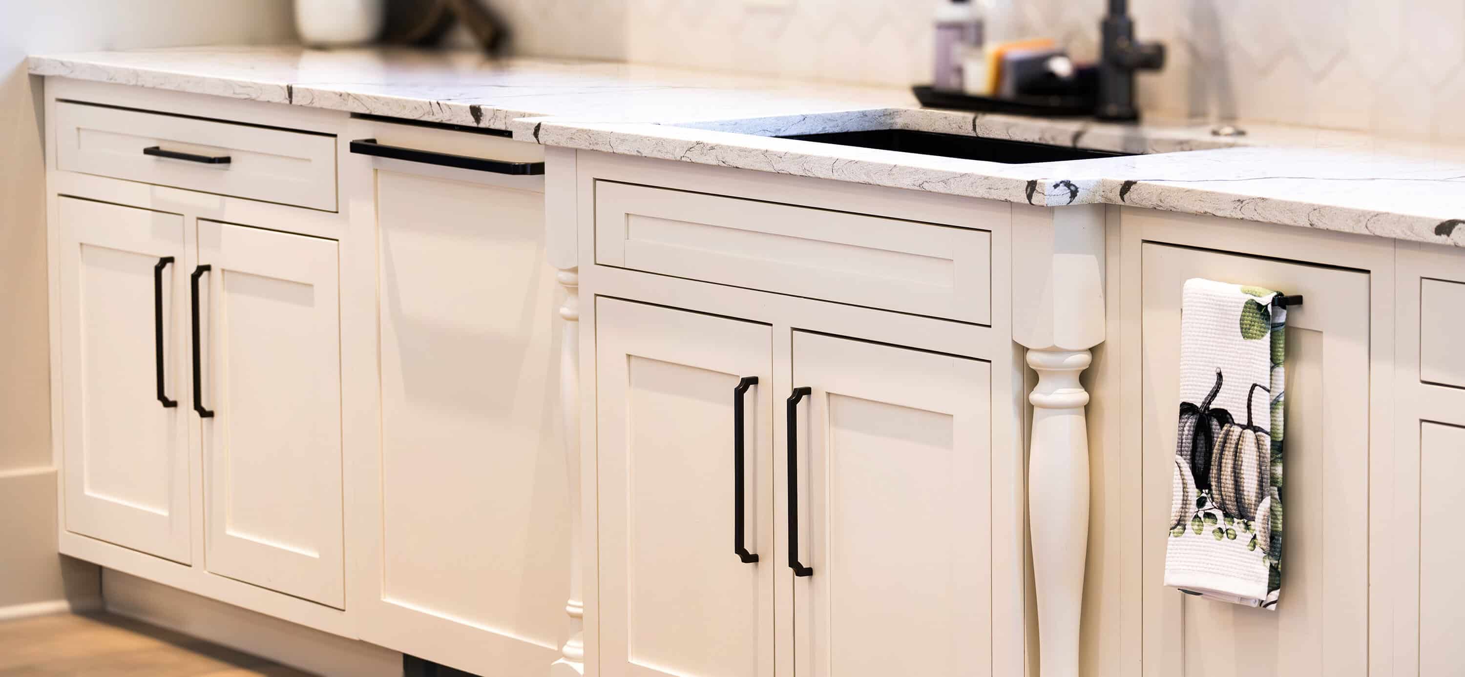 A sleek Knoxville kitchen showcases white cabinets with black handles. The natural stone countertop, adorned with a marble finish, supports a sink, and a dish towel with utensils hangs on the cabinet door. The background is softly blurred for an elegant touch.