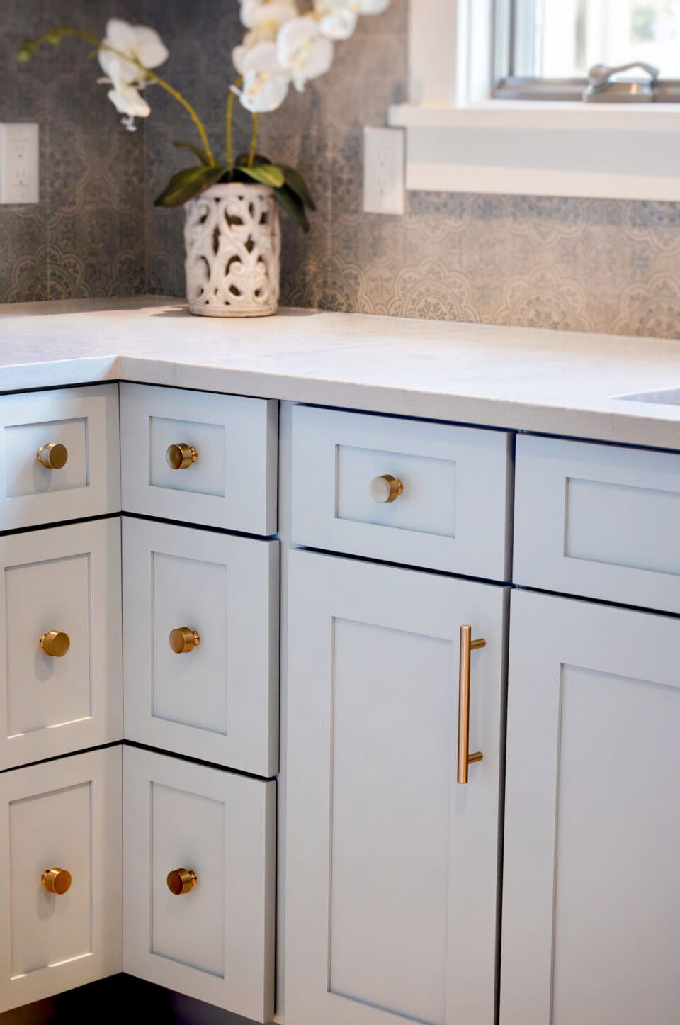 A modern kitchen corner in Knoxville features light gray cabinets with gold handles. The natural stone countertop gleams, accompanied by a vase of white orchids. Patterned gray tiles form the backsplash under a bright window, infusing the space with elegance and charm.