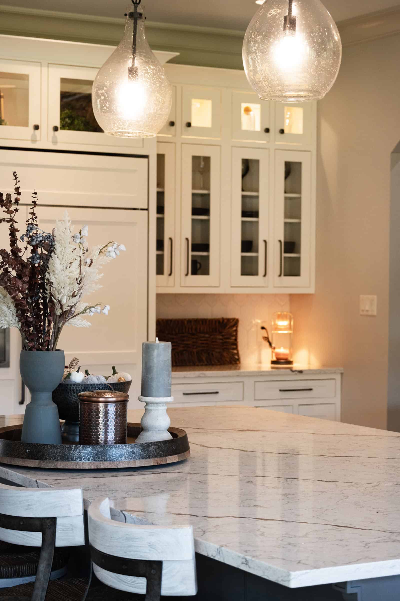 A modern kitchen with white cabinets, glass panel doors, and two pendant lights. The large marble island features a natural stone countertop adorned with a decorative tray holding vases and a candle. Wooden chairs with light cushions surround the island, while soft lighting creates a warm ambiance.