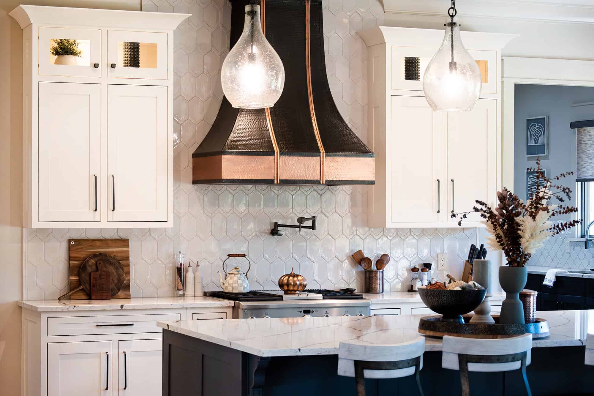 Modern kitchen in Knoxville with white cabinets, a large black exhaust hood with copper trim, and a marble island. Two pendant lights hang above the island, which has bar stools and a decorative centerpiece. The backsplash features hexagonal tiles and complements the natural stone countertop.