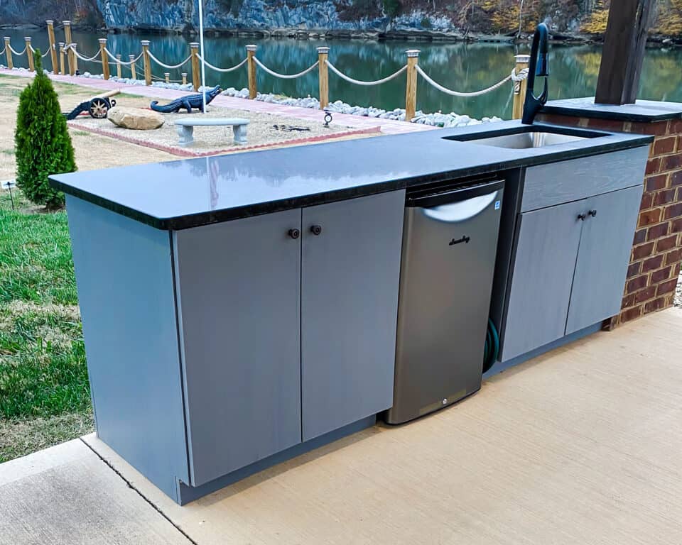 Outdoor kitchen setup with sleek gray cabinets and a modern black quartz countertop, featuring a built-in sink and mini refrigerator. The background showcases a serene lake, rope fence, and lush grassy area with shrubs on the stone-paved patio.