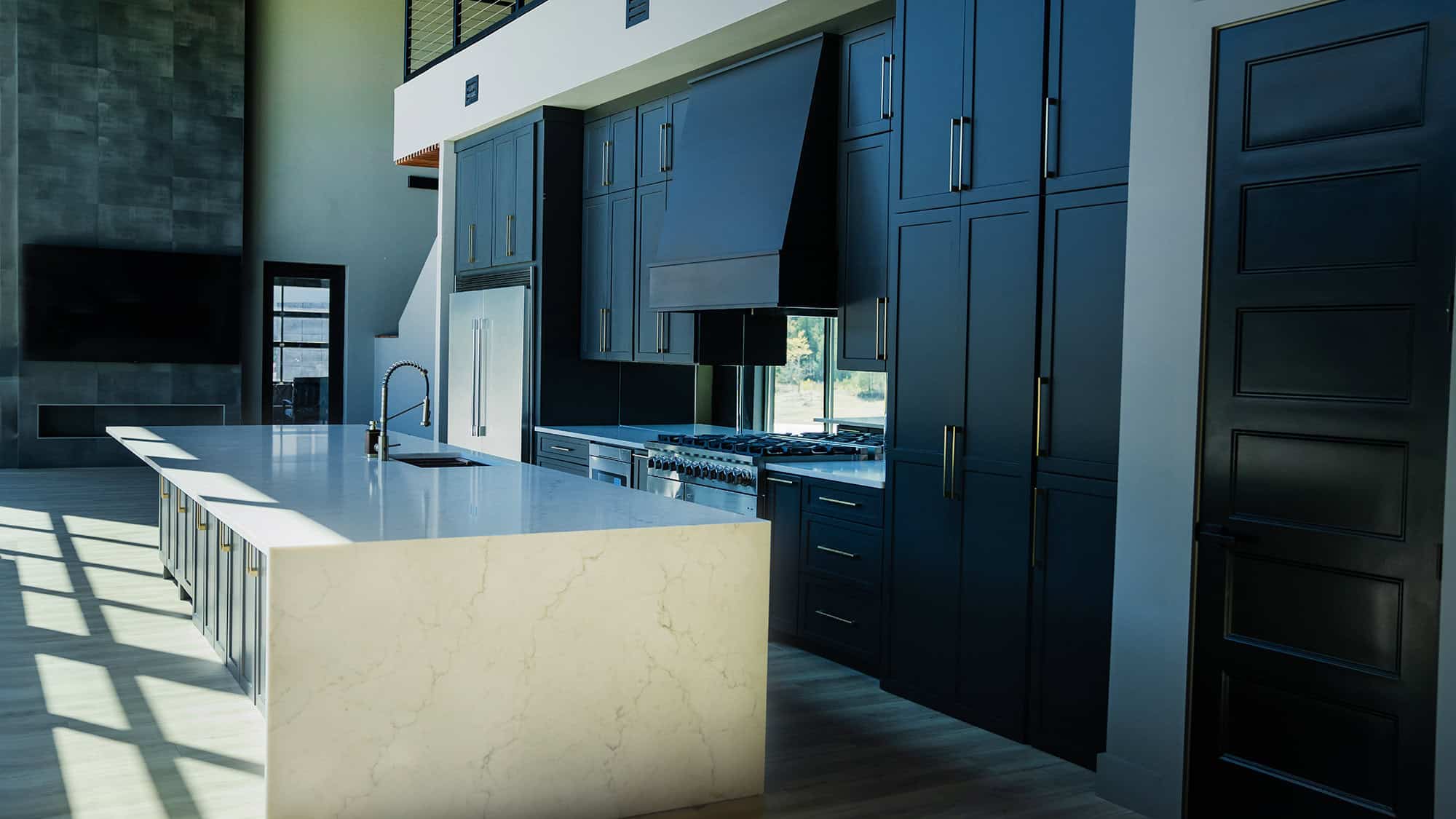 A modern kitchen in Knoxville boasts dark cabinets and a sleek marble island with a quartz countertop. The space features stainless steel appliances, a large sink, and a black range hood. Sunlight streams through tall windows, casting shadows on the wooden floor.