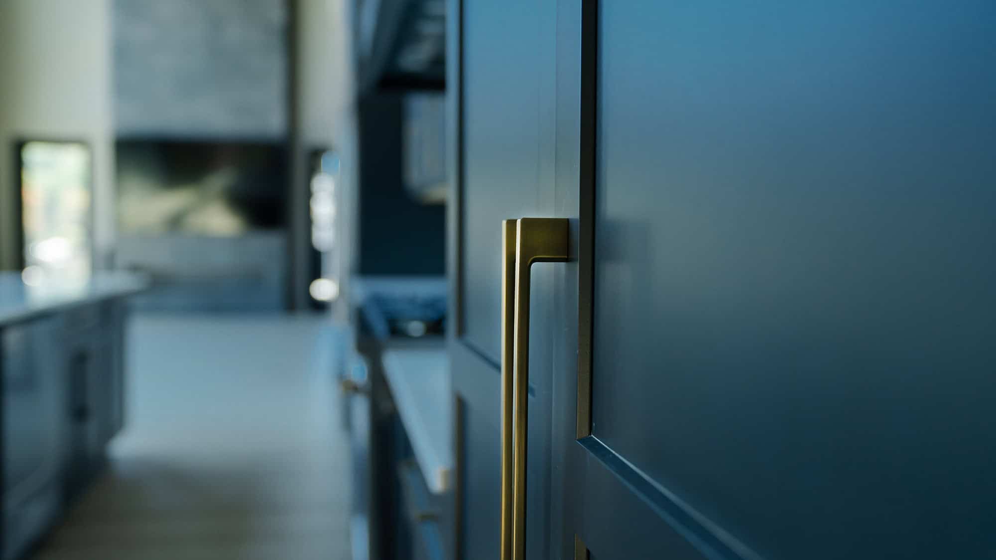 Close-up of a modern kitchen in Knoxville with blue cabinets and brushed metal handles. The background shows a blurred open-plan area with natural light coming from a window, highlighting sleek countertops and a minimalistic interior design.