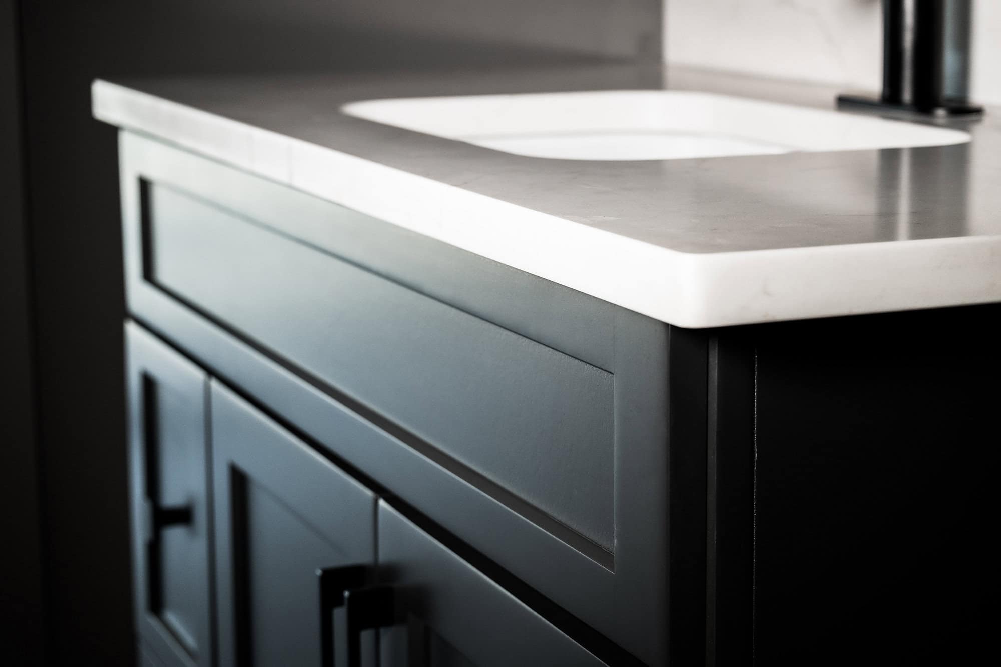 Close-up of a modern bathroom vanity in Knoxville, featuring a sleek quartz countertop and square sink. The dark cabinet boasts subtle paneling and dark handles, creating a striking contrast with the light surface above.