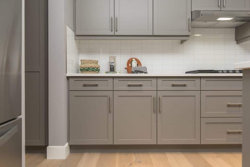 Modern kitchen with gray cabinets and stainless steel handles. The natural stone countertop features a cutting board, a set of woven baskets, and a storage canister. The backsplash is white tile, and the wood floor adds warmth to the space.