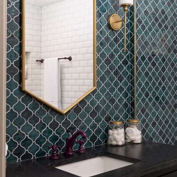 A bathroom with a natural stone countertop and a white sink, featuring teal and white patterned tiles on the wall. A geometric mirror hangs above, complemented by jars with cotton balls on the counter. Sleek cabinets add style, while a towel is visible on a rod in the background.